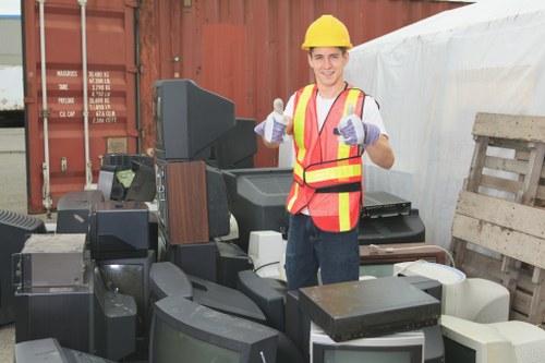 A male worker wearing a yellow safety helmet, high-visibility orange vest with yellow reflective strips, and purple work gloves stands among a collection of discarded electronic appliances and waste items. The appliances include old computer monitors, CRT screens, and various electronic devices stacked on a concrete or paved surface outdoors. In the background, there is a large rusted shipping container, hinting at a storage or recycling yard, with a white tarp and wooden pallets nearby. The scene appears to be part of a waste collection or clearance operation, possibly managed by Commercial Waste Removal Staines, with the worker giving a thumbs-up gesture indicating successful rubbish removal. The environment is well-lit with natural daylight, providing clear visibility of the dusty and weathered surfaces of the electronic waste and surrounding setting, aligning with local rubbish removal services in the Staines area.