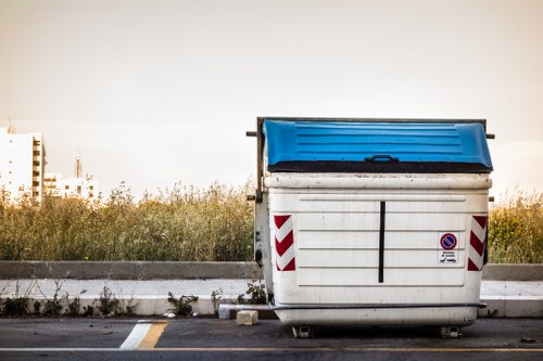 A large outdoor commercial waste container with a predominantly white body and red diagonal reflective stripes on the sides, positioned on a paved surface next to a curb. The container has a blue lid with a slightly textured surface, partially closed, and appears to be made of metal or durable plastic. It sits in a parking or roadside area with a low concrete curb and some grass and weeds growing along the edge of the pavement. The background features a clear sky and a distant view of an industrial or townscape with tall buildings or cranes, indicating a location that could be in or near Staines. The scene is evenly lit, suggesting natural daylight, and the container is associated with rubbish or waste removal services such as those offered by Commercial Waste Removal Staines, fitting within a typical urban or suburban environment where commercial waste is collected and managed.