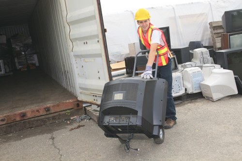 A female worker wearing a yellow safety helmet, high-visibility vest with red and yellow stripes, and white gloves is seen lifting a black office monitor with a removed stand, preparing for rubbish removal. She is standing on a paved outdoor surface near a large open shipping container, which reveals an interior filled with various discarded electronic components and boxes. The surrounding area includes a stack of old computer monitors, small cardboard boxes, and other miscellaneous waste stacked against the container wall, indicative of commercial or electronic waste clearing. The environment appears to be a waste disposal or recycling yard in Staines, with diffuse daylight illuminating the scene. The professional context aligns with rubbish removal services provided by Commercial Waste Removal Staines, emphasizing proper handling of electronic waste and bulk debris typical in commercial decluttering or disposal projects.