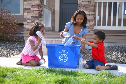 A woman and two children, a girl wearing a pink and white striped shirt with a pink skirt and a boy in a red shirt with dark shorts, are outdoors on a grassy front garden of a residential property, engaged in recycling activities. The woman, standing between the children, is holding a clear plastic bottle and placing it into a blue recycling bin marked with the recycling symbol and the words 'WE RECYCLE.' The girl is kneeling on the grass, holding a plastic bottle close to her mouth, while the boy sits on the paving beside the bin, clutching a plastic bottle in his hand. The background features a stone and brick exterior of the house with a dark grey front door and a white railing on the porch, indicating a suburban setting in Staines. The scene is brightly lit by natural daylight, enhancing the vibrant colours of the children's clothing and the green grass, illustrating a family participating in environmentally conscious waste management at their home in Surrey.