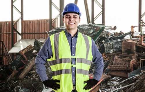 A young male waste management professional wearing a blue safety helmet and a high-visibility yellow vest over a dark shirt, standing inside a spacious industrial warehouse. Behind him, there is a large pile of various waste materials, including metal scrap, wooden debris, and broken appliances, scattered across the concrete floor. The background features tall steel framework and natural light filtering through large windows. The worker is smiling confidently with hands on hips, indicating a positive approach to rubbish removal services. This image reflects the type of waste collection and clearance work conducted by Commercial Waste Removal Staines, serving the Staines area with professional junk disposal in an industrial setting.