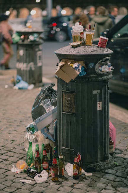 A black cylindrical public trash bin made of metal with vertical ridges and a slightly domed top, situated on a cobblestone street. The open lid reveals a few paper cups and snack wrappers on top of t