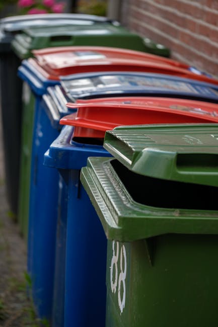 A black cylindrical public trash bin made of metal with vertical ridges and a slightly domed top, situated on a cobblestone street. The open lid reveals a few paper cups and snack wrappers on top of t